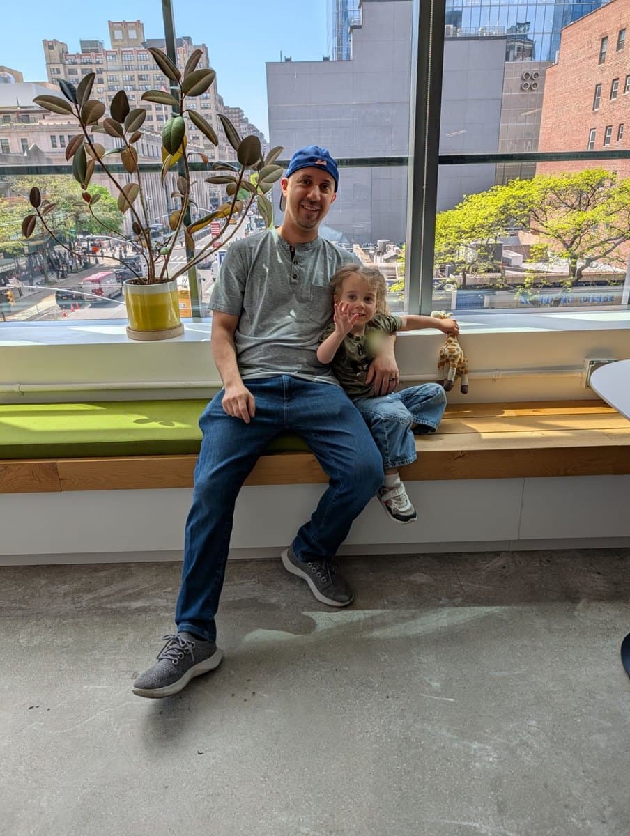 Keith, in a Mets cap, on a window bench with his daughter waving beside him and Manhattan behind them